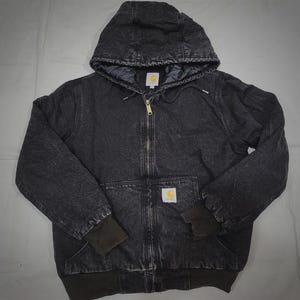 May include: A dark grey hooded jacket with a full-length zip and a small logo patch. The jacket features a quilted lining, ribbed cuffs, and two front pockets. It is made from a durable, workwear-style fabric.