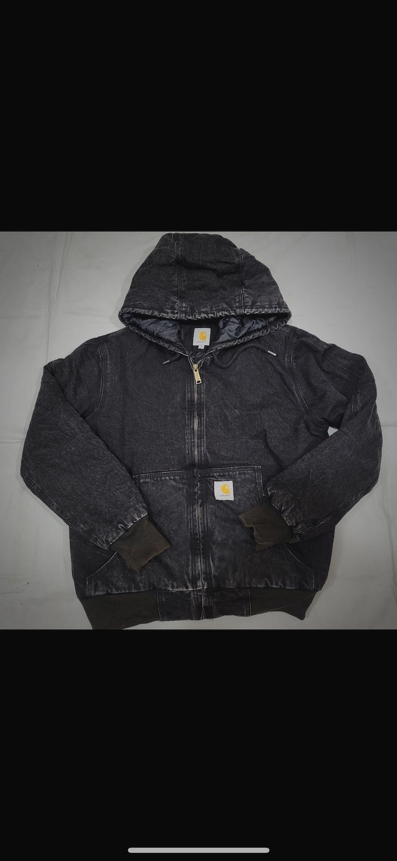 May include: A dark grey hooded jacket with a full-length zip and a small logo patch. The jacket features a quilted lining, ribbed cuffs, and two front pockets. It is made from a durable, workwear-style fabric.