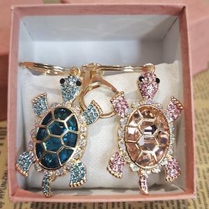 Puede incluir: Dos llaveros de tortuga enjoyados. Una tortuga tiene un caparazón verde azulado y detalles azules, la otra tiene un caparazón melocotón y detalles rosas. Ambos tienen marcos y llaveros de metal dorado. Se exhiben en una caja de regalo rosa.