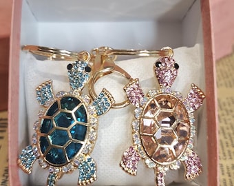 May include: Two bejeweled turtle keychains. One turtle has a teal shell and blue accents, the other has a peach shell and pink accents. Both have gold-colored metal frames and key rings. They are displayed in a pink gift box.