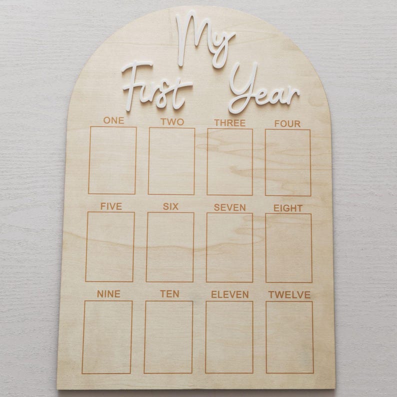 May include: Wooden milestone board with an arched top. The words 'My First Year' are in white, cursive lettering. Below are twelve rectangular spaces labeled with the months 'ONE' through 'TWELVE'. The board is made of light-colored wood with visible grain.