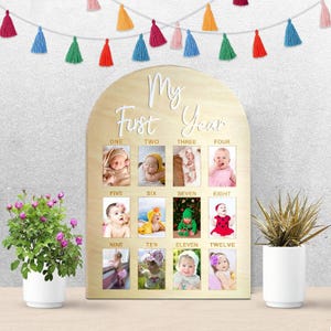May include: Wooden baby milestone photo frame with a light brown finish. The frame has an arched top with the words 'My First Year' in white script. It features twelve photo slots labeled with the months 'ONE' through 'TWELVE'. The background is a light gray. A garland of colorful tassels hangs above the frame. Two potted plants are on either side of the frame.