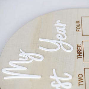 May include: A wooden milestone disc with white raised lettering. The disc is round and has the words 'My first year' in a cursive font. The right side of the disc has boxes with the words 'TWO', 'THREE', and 'FOUR' printed vertically. The wood has a light, natural color.
