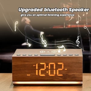 May include: A wooden alarm clock with a digital display showing 12:02 AM. The clock has a built-in Bluetooth speaker and a wireless charging pad. The clock is on a wooden surface with a blurred background.
