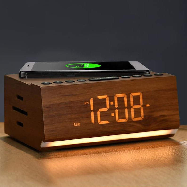 May include: A wooden alarm clock with a digital display showing 12:08 AM. The clock has a built-in wireless charging pad for a smartphone, which is shown on top of the clock. The clock has a light brown finish and a white light illuminating the display.