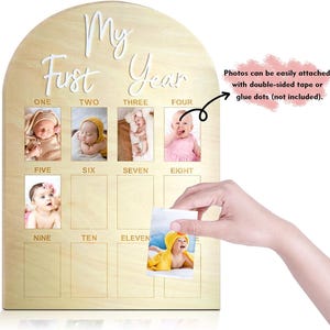 May include: Wooden baby monthly milestone photo board. The board is arched at the top and has the words 'My First Year' in white script. It has spaces for twelve photos, labeled with the months one through eleven. The board includes a note that photos can be attached with double-sided tape or glue dots.