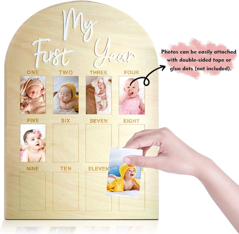 May include: Wooden baby monthly milestone photo board. The board is arched at the top and has the words 'My First Year' in white script. It has spaces for twelve photos, labeled with the months one through eleven. The board includes a note that photos can be attached with double-sided tape or glue dots.