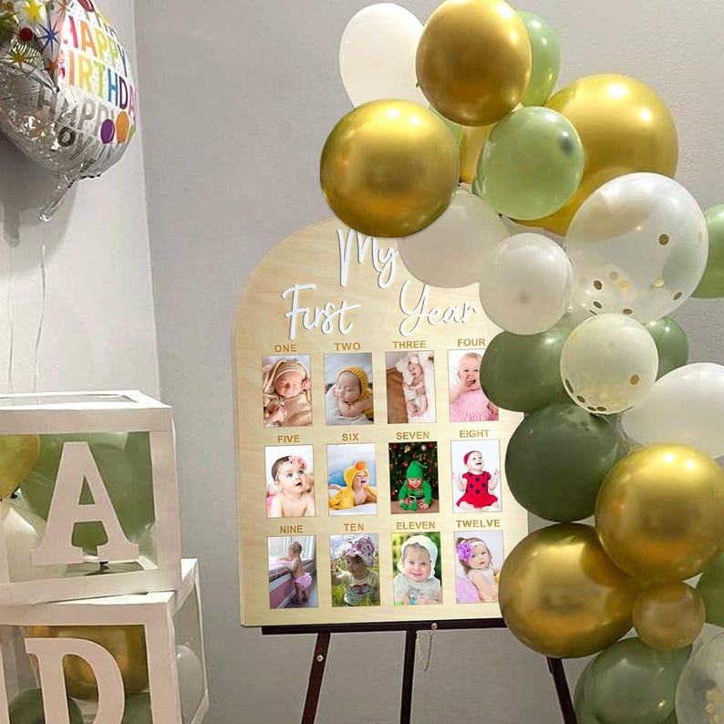 May include: A wooden sign with photos of a baby's first year. The sign has a light wood finish and an arch shape. It features twelve small photo frames, each labeled with a month from one to twelve. The sign is decorated with gold, white, and green balloons, and a balloon that says 'Happy Birthday'.