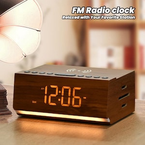 May include: A brown wooden digital clock radio with a large orange digital display showing the time 12:06 AM. The clock has a built-in FM radio and a wireless charging pad. The clock is on a wooden surface.