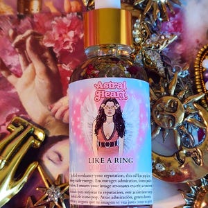 May include: A glass bottle of Astral Heart oil with a gold cap and dropper. The label features an illustration of a woman with angel wings and the text "LIKE A RING". The background includes gold celestial decorations.