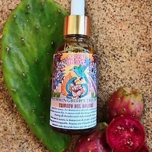 May include: A glass bottle with a gold cap and a colorful label featuring a hummingbird and text. The bottle is surrounded by a green cactus pad and several red prickly pears. The label reads "Hummingbird's Triumph".