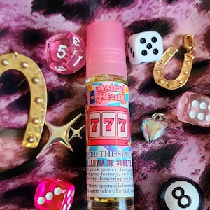 Puede incluir: Un frasco de vidrio transparente con tapa rosa y una etiqueta con el texto Astral Heart, 777 y Roll the Seven. El frasco está rodeado de varios dijes y piezas de juego, incluidos dados, una herradura, un corazón y una bola ocho.