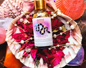 Può includere: Una bottiglia di vetro con un tappo dorato e un'etichetta rosa e viola che dice "Astral Heart Femme Fatale Mujer Fatal". La bottiglia è circondata da fiori rosa e bianchi e perle.