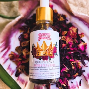 May include: A glass bottle with a gold cap and label. The label is pink and white with the text "Astral Heart Queens Envy Envidia de la Reina" and a crown with red roses. The bottle is sitting on a white seashell with pink and red flower petals.