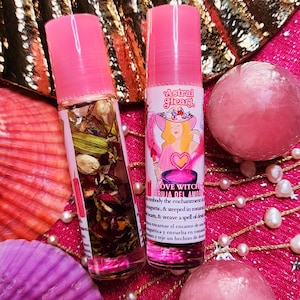 Może przedstawiać: Dwie różowe butelki z kulką z różowymi zakrętkami, jedna z suszonymi ziołami, a druga z etykietą "LOVE WITCH BRUJA DEL AMOR". Butelki znajdują się na różowej powierzchni z perłami i różowymi elementami dekoracyjnymi.
