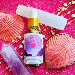 Könnte beinhalten: Eine Glasflasche mit goldener Kappe und einem Etikett mit der Aufschrift "Astral Heart 13 Moons Luna". Die Flasche steht auf einer rosa Oberfläche mit einem weißen Kristall, einer rosa Muschel und einem violetten Kristall.