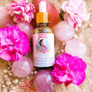 May include: A glass bottle with a gold cap and a pink label that says "Astral Heart" and "Siren Song Canto de Sirena". The bottle is surrounded by pink flowers, seashells, and pink and white stones.