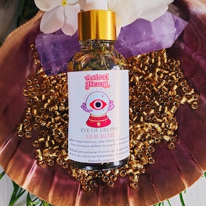 May include: A glass bottle of oil with a gold dropper, labeled "Eye of Delphi" with a crystal ball graphic. The bottle sits on a decorative shell with gold beads, white flowers, and a purple crystal.