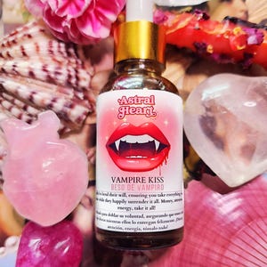 May include: A glass bottle with a gold cap and a red and white label. The label says "Vampire Kiss Beso de Vampiro" and has a cartoon vampire mouth with fangs. The bottle is sitting on a pink and white surface.
