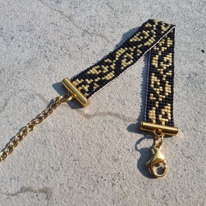 May include: A beaded bracelet with a black and gold geometric pattern. The bracelet has gold-colored metal end caps, a chain, and a lobster clasp. The bracelet is lying on a light gray surface.