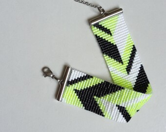 Puede incluir: Una pulsera de cuentas con un patrón geométrico en negro, blanco y verde neón. Tiene un cierre plateado y una cadena con un dije de estrella. La pulsera mide aproximadamente 2 cm de ancho.