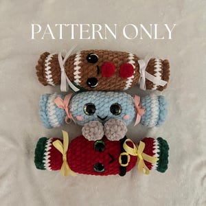 May include: Three crocheted Christmas cracker ornaments. The top cracker is brown and white with a red pom-pom. The middle cracker is blue and white with a gray pom-pom. The bottom cracker is red and green with a yellow bow.