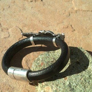 May include: A black leather bracelet with a silver clasp and a silver charm in the shape of a bird. The bracelet is lying on a brown surface.