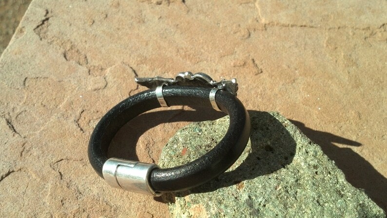 May include: A black leather bracelet with a silver clasp and a silver charm in the shape of a bird. The bracelet is lying on a brown surface.