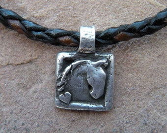 Op de afbeelding: Een zilveren ketting met een gevlochten leren koord. De hanger is een vierkant met een paardenkopontwerp en een klein hartje.