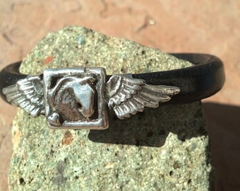 Op de afbeelding: Een zwart leren armband met een zilveren gevleugelde paardenbedel. De bedel is gedetailleerd met een paardenkop en vleugels.