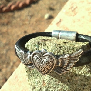 May include: A black leather bracelet with a silver heart charm featuring wings. The heart has a decorative design.