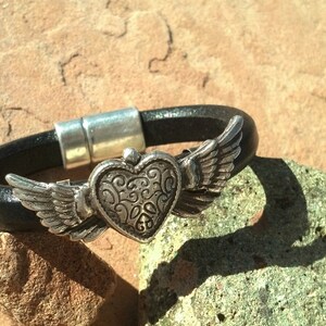 May include: A black leather bracelet with a silver heart charm with wings. The heart has a detailed design and the wings are spread out.