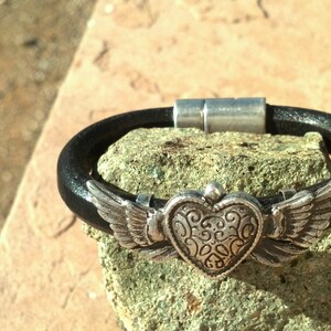 May include: A black leather bracelet with a silver heart charm with wings. The heart has a detailed design and the wings are spread out.