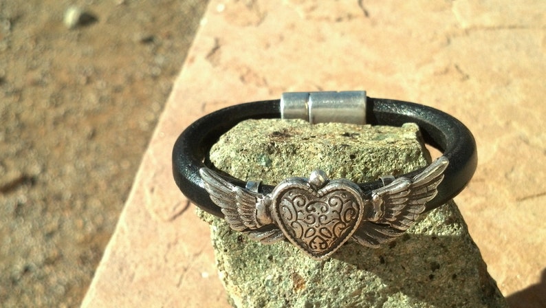 May include: A black leather bracelet with a silver heart charm with wings. The heart has a detailed design and the wings are spread out.