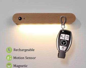 May include: A wooden LED light fixture with a key fob attached. The light is rectangular with rounded edges. The key fob is black and silver. The light is labelled as rechargeable, motion sensor, and magnetic. The light emits a warm glow.