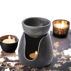 Pode incluir: Um difusor de óleos essenciais de cerâmica cinza com uma tigela redonda no topo e uma base em forma de gota. Uma vela de chá acesa fica dentro da base, e duas outras velas estão no fundo. As velas estão acesas, criando um ambiente acolhedor.