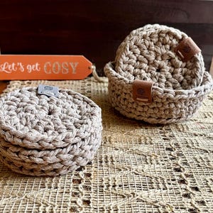May include: A set of round, beige crocheted coasters stacked in a matching crocheted basket. Each coaster and the basket features a small, rectangular brown leather tag with a yarn ball icon. An orange tag with the text Let's get COSY is visible.