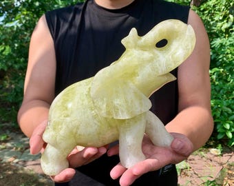 May include: A carved elephant figurine in shades of pale yellow and white. The sculpture is held in a person's hands, showcasing the detailed craftsmanship. The elephant has its trunk raised and ears flared, suggesting movement.