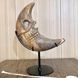 May include: A carved stone crescent moon sculpture with a skull-like face, supported by a black metal stand. The sculpture has a mix of tan, brown, and gray tones. The stand has a circular base.