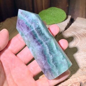 May include: A green, purple, and white fluorite crystal point. The crystal is smooth and polished, with a pointed tip and a flat base. It is held in a hand, and the background is a wooden surface.