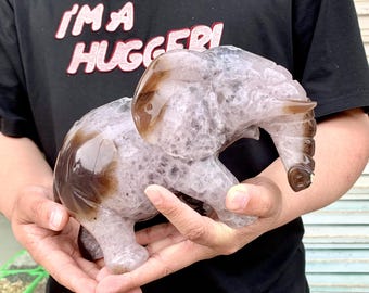 May include: A carved elephant figurine in shades of brown and grey is held in two hands. The elephant has a detailed trunk, ears, and body. The figurine is made of a stone-like material and has a polished appearance.