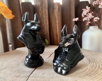 May include: Two black obsidian Anubis statues, one with a more detailed design than the other, are displayed on a wooden surface.