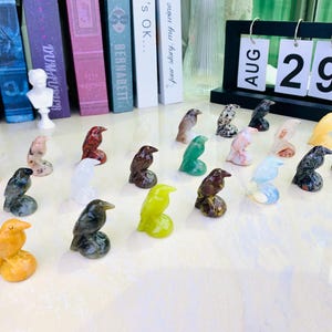 May include: A selection of carved stone bird ornaments in diverse colours, such as green, yellow, and brown. The birds are on small bases. A calendar in the background shows "AUG 29".