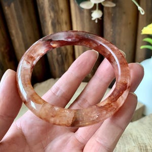 May include: A translucent, reddish-brown gemstone bangle bracelet. The bracelet is a solid, circular band with a polished surface. The colour is a mix of clear and opaque tones, with streaks of red and brown. The bracelet is held in a hand.