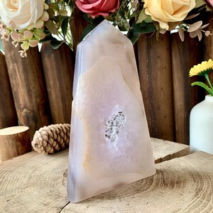 Può includere: Un grande cristallo di agata lucidata nei toni del bianco, crema e grigio, con un centro di geode. Il cristallo è esposto su una superficie di legno, con fiori e una pigna sullo sfondo. Il cristallo è alto circa 20 cm.