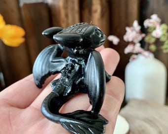 May include: A black figurine of Toothless, the Night Fury dragon from How to Train Your Dragon, with a detailed textured body and wings. The dragon is posed with its head tilted up and its tail curled behind it.