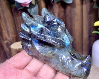 May include: A carved labradorite dragon head sculpture. The dragon head is primarily gray with iridescent blue and green flashes. The sculpture has detailed features, including horns and a snout. The dragon is held in a hand.