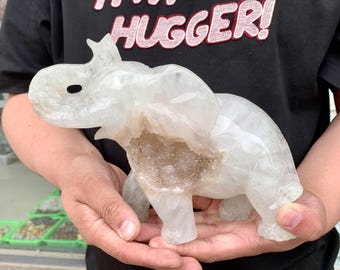May include: A carved, translucent quartz elephant figurine. The sculpture features a hollowed-out section revealing a crystal-lined interior. The elephant is held in open hands, showcasing its detailed form and natural colour variations. The figurine is a decorative item.