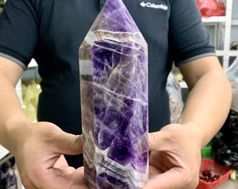May include: A large, polished amethyst crystal point. The crystal features a deep purple hue with white and translucent inclusions. The top is a pointed pyramid shape. The crystal is held in a person's hands.