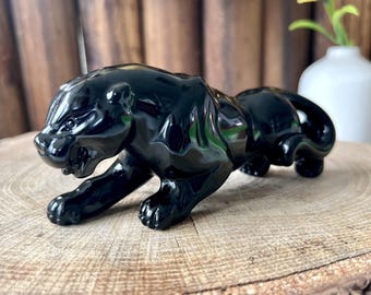 May include: A glossy black panther figurine in a stalking pose. The sculpture is set on a natural wood slice, with a blurred background featuring flowers and a wooden wall. The panther's sleek form and detailed features are highlighted.